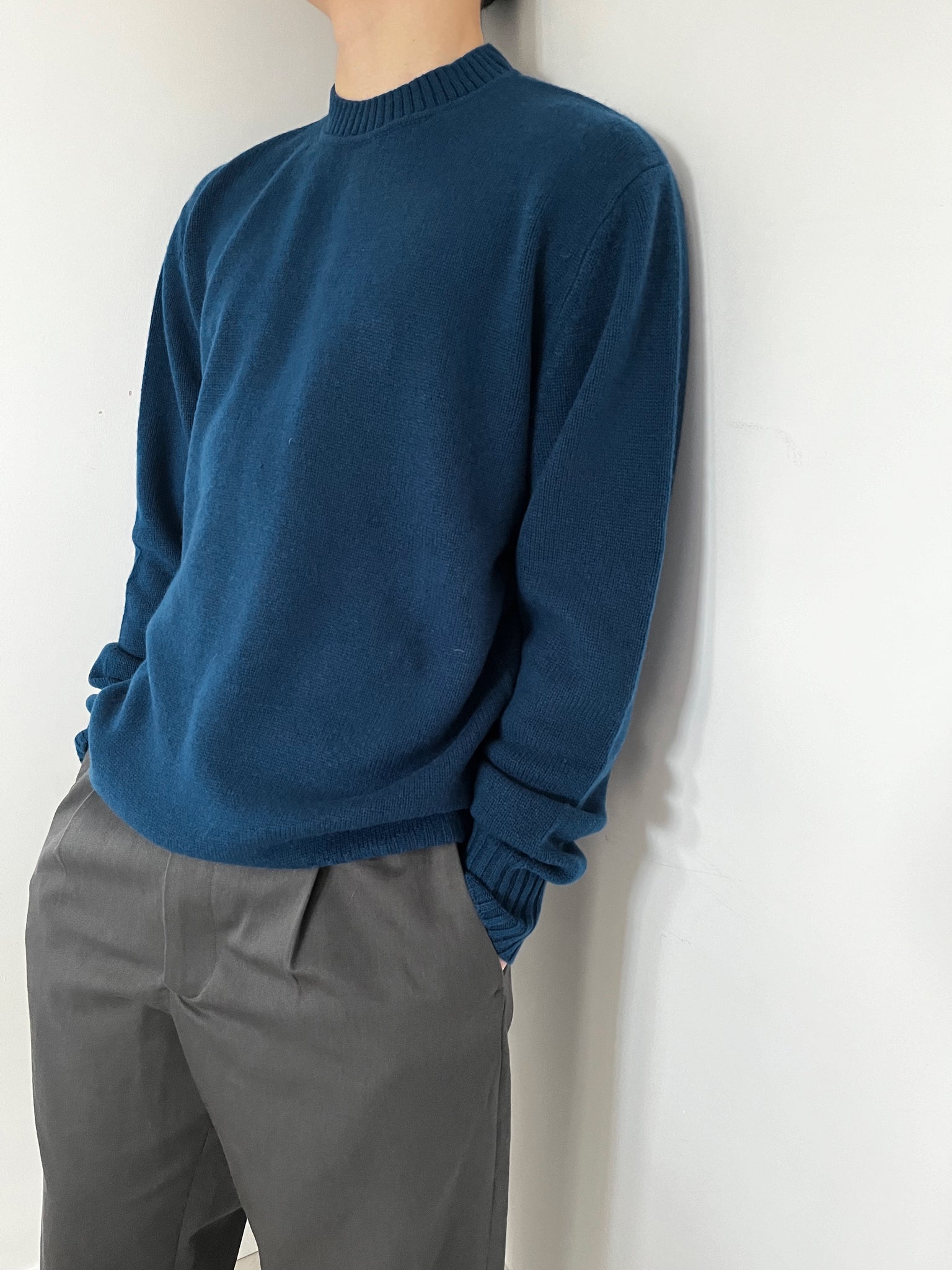 Person wearing a blue sweater and grey pants against a white wall